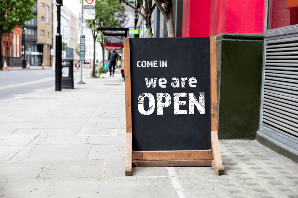 A clear outdoor sign stating "We Are Open," positioned on the sidewalk to attract potential customers.